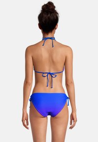 Vrouw met donker haar in een knot, gekleed in een blauwe en paarse gradient bikini onderkant en een blauwe halterbikini-top, gezien vanuit de rug.