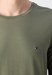 Oliwkowa, długosmykowa koszula wykonana z miękkiego materiału, z okrągłym dekoltem i małym haftowanym logo w kolorach czerwonym, białym i niebieskim.