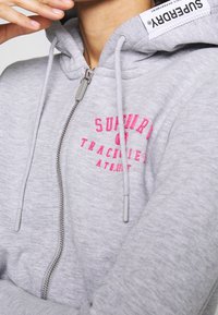 Sudadera con capucha gris con cremallera, texto bordado en rosa, cremallera plateada y cordones grises. Cuenta con una capucha forrada y una textura suave de algodón.