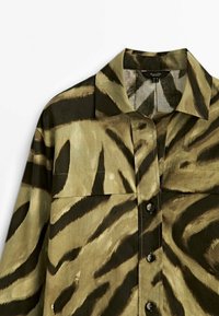 Olive green shirt with black tiger stripe pattern, featuring a collar, button-down front, and two chest pockets. Textured fabric.