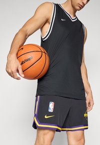 Top de basquetebol sem mangas preto com detalhes brancos e o logo da Nike, combinado com calções pretos com detalhes em roxo e amarelo, segurando uma bola de basquetebol laranja.