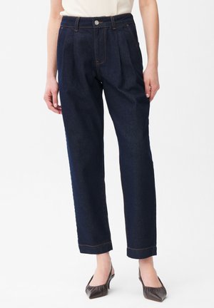 Donna che indossa jeans plissettati a vita alta di colore blu scuro e tacchi scuri con punta appuntita, in piedi su uno sfondo bianco.