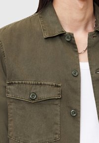 Olivegrünes Hemd aus Denimstoff mit Knöpfen. Es verfügt über einen Kragen, eine Brusttasche mit Knopf und eine strukturierte Oberfläche.