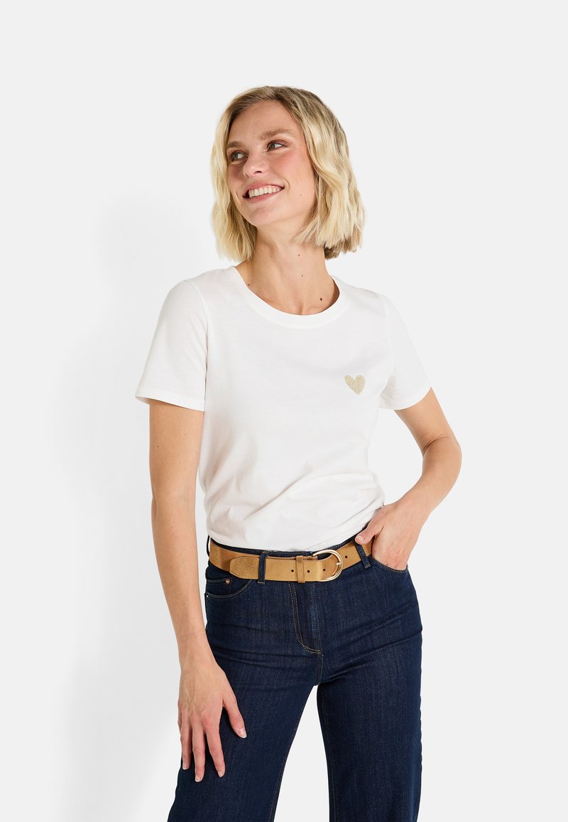 Camiseta blanca de algodón con cuello redondo y mangas cortas, con un pequeño diseño de corazón dorado en el pecho, combinada con jeans azul oscuro y un cinturón marrón claro.