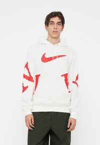 Bela kapuca s kapuco, ki ima velike rdeče Nike logotipe spredaj in na rokavih. Material se zdi mehak, s kenguru žepom in rebrastimi manšetami.