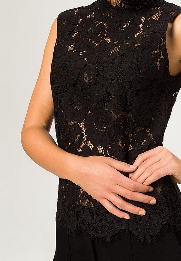 Haut en dentelle noire sans manches avec motif floral, col montant et ourlet festonné, porté par une personne avec les mains doucement jointes à la taille.