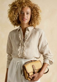 Svetlo béžová pruhovaná košeľa s oblúkovým golierom, v kombinácii s bielou sukňou na šnúrku. Drží tkanú prútenú kabelku s hnedými akcentmi.