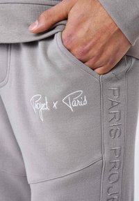 Joggers gris avec un logo brodé "Project x Paris" sur le côté et une poche avant. Le tissu semble doux et lisse.