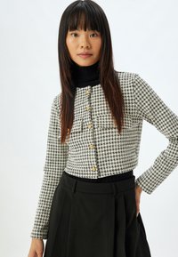 Schwarze und weiße Hahnentrittmuster-Cropped-Jacke mit langen Ärmeln, goldenen Knöpfen und einem steifen Kragen. Kombiniert mit einem schwarzen, plissierten Rock.