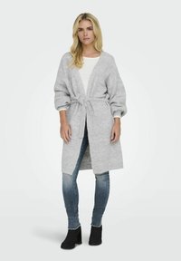 Cardigan long gris clair avec une taille à nouer, texture côtelée et manches larges. Associé à un haut blanc et un jean, porté avec des bottines noires.
