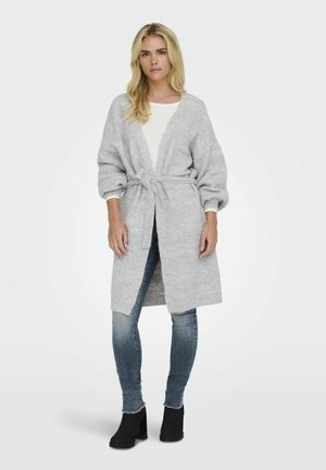 Cardigan lungo grigio chiaro con cintura in vita, texture a coste e maniche larghe. Abbinato a un top bianco e jeans, indossato con stivali neri alla caviglia.