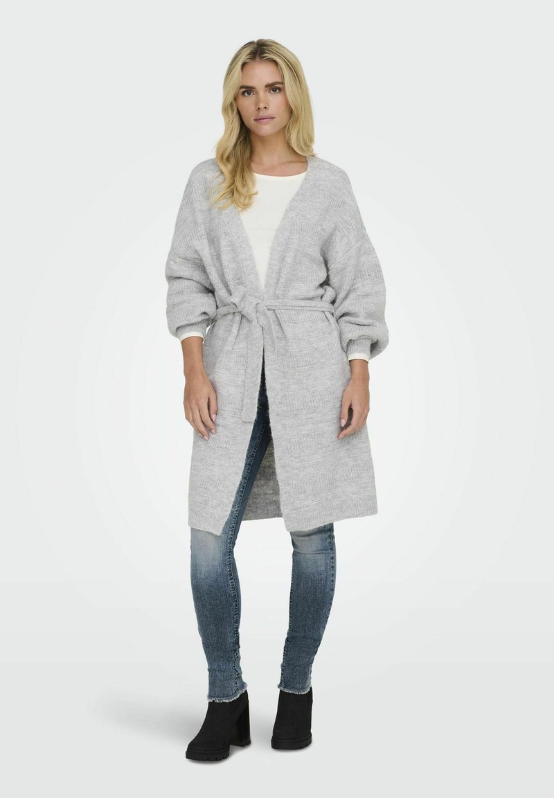 Cardigan long gris clair avec une taille à nouer, texture côtelée et manches larges. Associé à un haut blanc et un jean, porté avec des bottines noires.