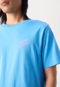 Světle modré tričko s krátkým rukávem vyrobené z bavlny, s růžovým grafickým potiskem a textem "SCOTCH & SODA" na levé přední straně.