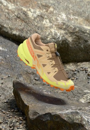 Salomon Trailrunning-Schuh mit braunem Obermaterial, hellgelber Sohle mit orangen Akzenten, strukturierten Materialien und leichtem Design.