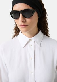 Czarne okrągłe okulary przeciwsłoneczne z ciemnymi szkłami, błyszczącymi oprawkami. Noszone z białą koszulą z kołnierzykiem z button-down. Widoczna czarna opaska na włosy.