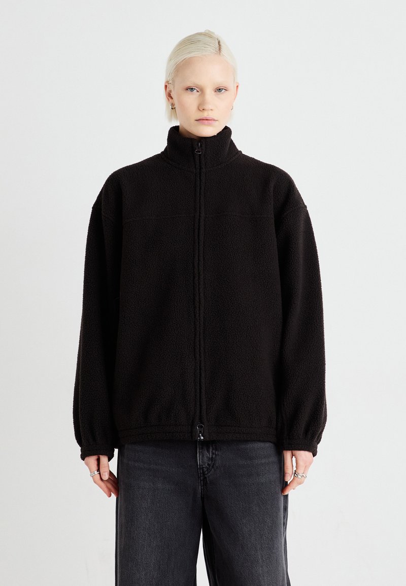 Schwarze Fleecejacke mit hohem Kragen, Reißverschluss und elastischen Bündchen. Hat eine strukturierte Oberfläche und einen lockeren Schnitt. Getragen mit dunklen Hosen.