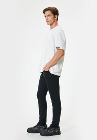 T-shirt bianca oversize con maniche corte, etichetta sulla manica, abbinata a jeans neri slim-fit e scarpe da ginnastica nere con fantasia.