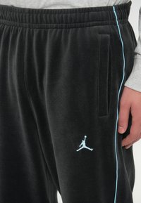 Juodos fliso sportinės kelnės su elastingu juosmeniu, šoninėmis kišenėmis ir šviesiai mėlynomis juostelėmis. Kairėje šlaunies pusėje yra baltas Jordan logotipas.