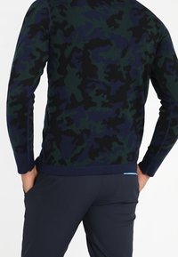 Homme portant un pull à manches longues, de couleur bleu foncé et vert avec un motif camouflage, les mains dans les poches du pantalon bleu marine, vu de dos.