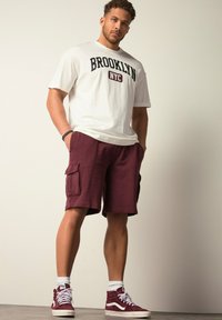 Camiseta blanca con la impresión "BROOKLYN NYC", pantalones cortos cargo burdeos con bolsillos laterales, calcetines blancos hasta el tobillo y zapatillas de deporte burdeos altas.