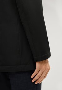 Schwarze Jacke aus strukturiertem Stoff, mit einer klaren Schnittführung, sichtbaren Nahtdetails und einem umgeschlagenen Ärmel. Hand liegt nahe dem Saum.