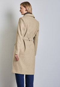 Beżowy płaszcz z wełny z domieszką, z kołnierzem, z paskiem w talii i prostym krojem. Widoczne boczne kieszenie i dopasowane szwy.