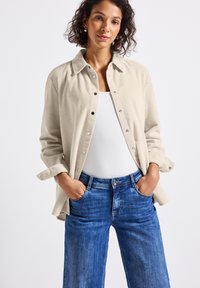 Vrouw met krullend haar, gekleed in een beige corduroy overhemd over een wit shirt en blauwe jeans, met handen in de zakken, staand tegen een effen achtergrond.