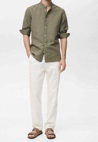 Olivengrünes Leinenhemd mit Stehkragen und Knopfleiste; kombiniert mit weißen Hosen und braunen Sandalen, die eine lockere Passform präsentieren.