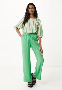 Groene wijde broek met elastische tailleband, gecombineerd met een gestreepte, lichtgekleurde blouse met knopen. De blouse heeft pofmouwen.