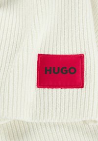 Maglione lavorato a maglia color crema con texture a costine. Presenta un'etichetta rettangolare rossa prominente con la scritta "HUGO" in lettere nere.