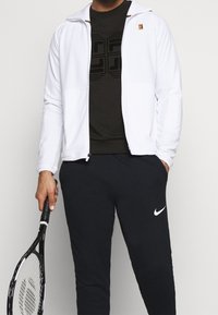 Biała kurtka zapinana na zamek na czarnym t-shircie z teksturowanym wzorem, w połączeniu z czarnymi spodniami dresowymi z białym logo Nike, trzymająca rakietę tenisową.