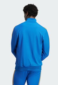 Blå fleece træningsjakke med høj krave, ribbede manchetter og kontrasterende orange side striber, vist fra bagsiden.