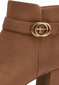 Brown suede ankle boot featuring a gold circular buckle detail and a block heel, with a smooth texture and stitched accents.