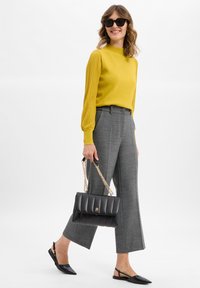 Gelber Pullover mit gerippten Ärmeln, graue plissierte Hose, schwarze gesteppte Handtasche mit goldener Kette und schwarze spitze Ballerinas.