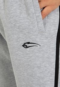 Hand in der Tasche einer hellgrauen Jogginghose mit schwarzem Streifen an der Seite und kleinem schwarzen gestickten Tierlogo nahe der Tasche.