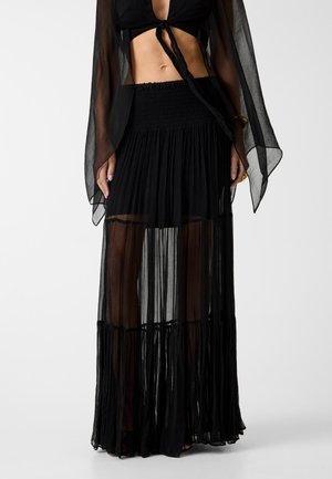 Femme portant une jupe maxi noire transparente avec une taille smockée et un haut court noir avec de longues manches transparentes, posant devant un fond uni.
