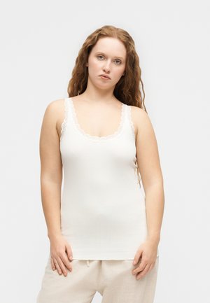 Femme aux longs cheveux bouclés portant un débardeur blanc orné de dentelle et un pantalon beige, debout devant un fond clair uni.