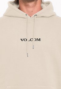 Hoodie beige avec une poche kangourou avant, des cordons de serrage et un logo "VOLCOM" brodé en noir centré sur la poitrine. Texture en coton lisse.