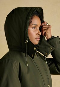 Oliwkowa, rozpinana kurtka z kapturem, wykonana z gładkiego materiału, z złotymi akcentami, regulowanymi sznurkami ściągającymi oraz przednim zapięciem na napy.