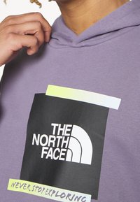 Hoodie de algodón morado con un gran gráfico negro que presenta "THE NORTH FACE" y "NEVER STOP EXPLORING" en acentos en blanco y verde neón.