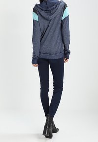 Marinblå hoodie med ljusblå och svarta axelinslag, avslappnad passform, matchad med mörka jeans och ankelskor. Mjuk textur, avslappnad design.