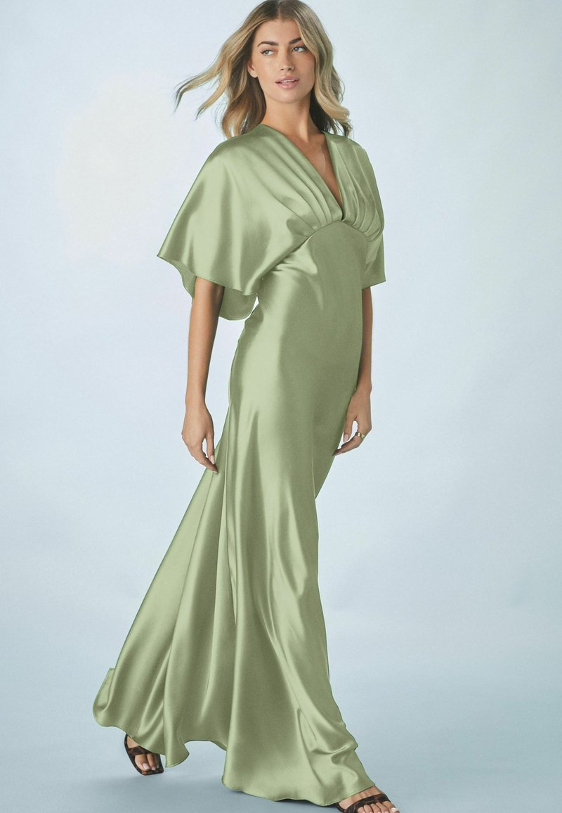 Satin grön klänning med djup V-ringning, draperade ärmar och en flödande kjol. Slät textur med subtil lyster, kombinerad med svarta sandaler med remmar.