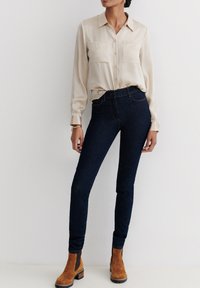 Beige Langarm-Hemd mit Knöpfen und zwei Brusttaschen, kombiniert mit dunkelblauen skinny Jeans und hellbraunen Stiefeletten. Glattes Material, taillierte Passform.