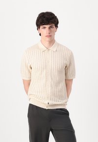 Polo din tricot bej, cu mânecă scurtă, cu un model texturat, guler ascuțit și închidere cu nasturi pe față. Material neted și design ajustat.