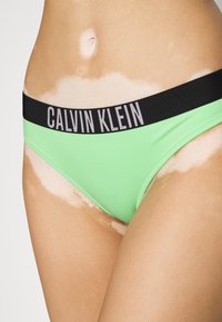 Šviesiai žalias bikini, su juodu elastingu juosmeniu, kuriame yra sidabrinėmisRaidėmis užrašyta "CALVIN KLEIN". Lygus paviršius, minimalios aprangos dizainas.