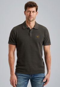 Mörk olivgrön polo-shirt med en texturerad väv, korta ärmar, krage och en liten guldig etikett på bröstet. Bärs med blå jeans.