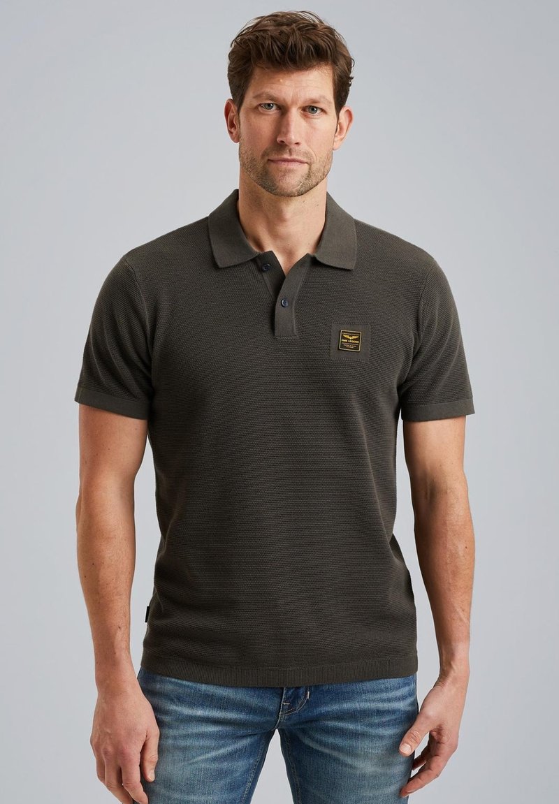 Mörk olivgrön polo-shirt med en texturerad väv, korta ärmar, krage och en liten guldig etikett på bröstet. Bärs med blå jeans.