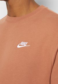 Brun bomullströja med rund halsringning, ribbad nederkant och en vit broderad Nike-logga på vänster bröstområde.