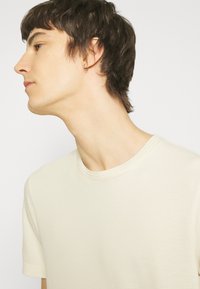 Kortærmet, cremefarvet T-shirt med et struktureret, vaffelmønstret stof og rund halsudskæring. Viser en person i profil med ørering.