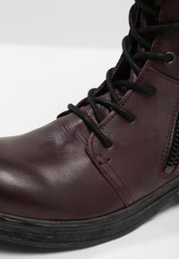 Stivale in pelle bordeaux con lacci neri e cerniera laterale, caratterizzato da una superficie leggermente graffiata e una suola in gomma nera.